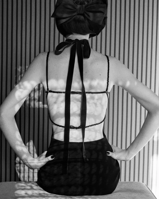 Back view of a woman with a large ribbon bow in her hair, wearing a black dress with spaghetti straps, against a striped wallpaper background.