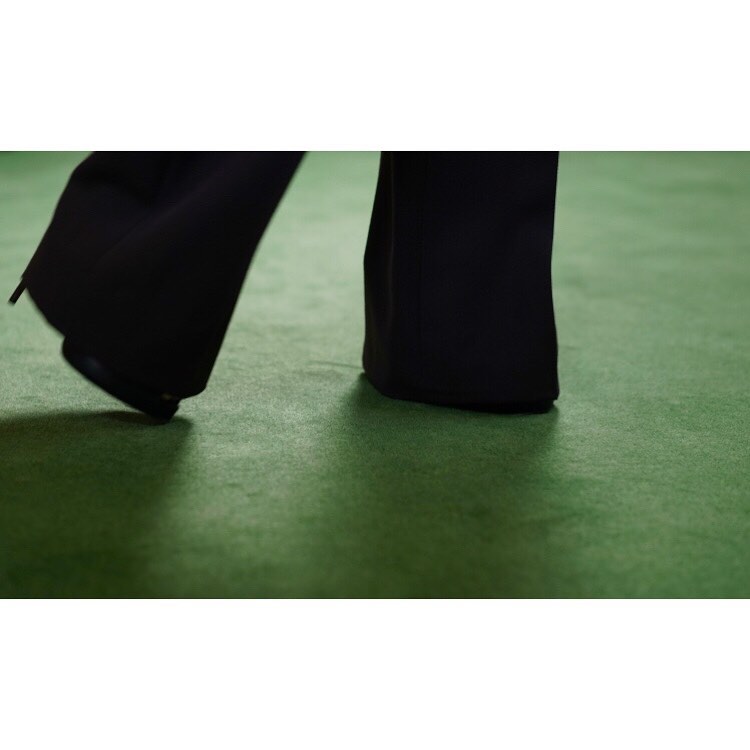Lower section of a person in black dress pants stepping onto a green carpet in a studio setting, captured at eye-level.