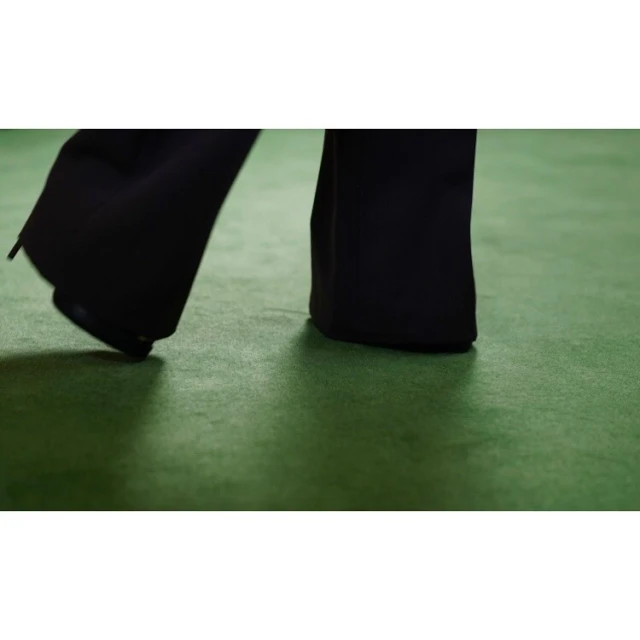 Lower section of a person in black dress pants stepping onto a green carpet in a studio setting, captured at eye-level.