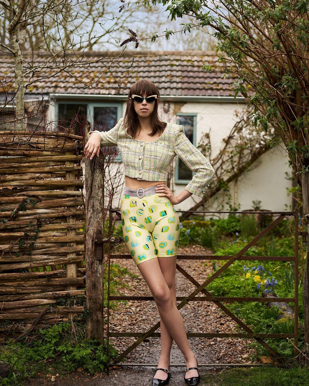 A young adult woman poses outdoors wearing a yellow cropped top and patterned shorts in front of a weathered fence and cottage.
