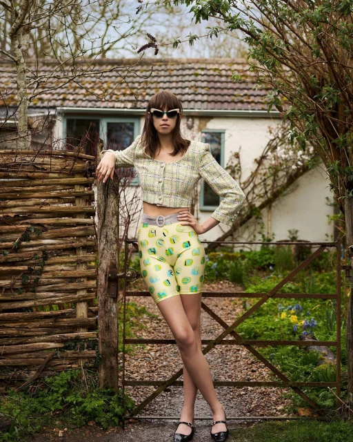 A young adult woman poses outdoors wearing a yellow cropped top and patterned shorts in front of a weathered fence and cottage.
