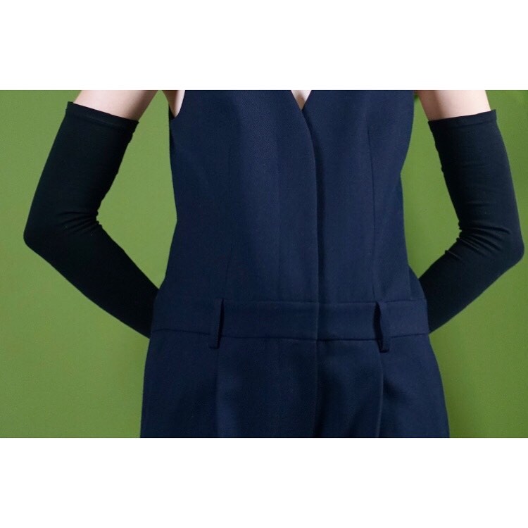 Woman in navy jumpsuit wearing black long-sleeved gloves poses against a light green background in a studio shot.