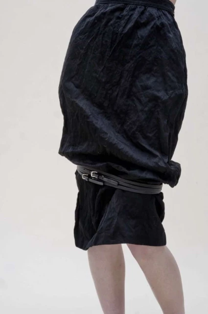 Side view of a person wearing a draped black skirt cinched with a black belt, legs visible from the knees down, against a plain background.