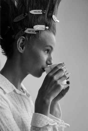 Monochrome portrait of woman drinking from paper cup with hair clips, side view, indoors against a plain wall, natural light.