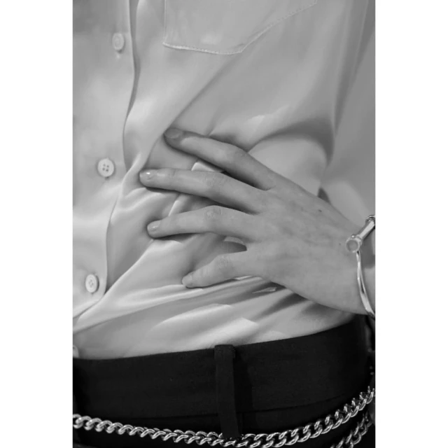 Close-up shot of a person in a white satin shirt, black trousers, and a silver chain belt and bracelet, set against a neutral background.