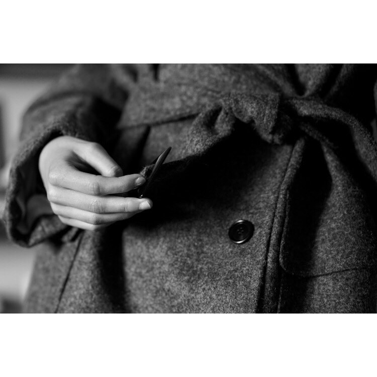 Close-up of a woman wearing a gray wool coat, fastening the tie. The black and white photo captures the texture of the fabric.