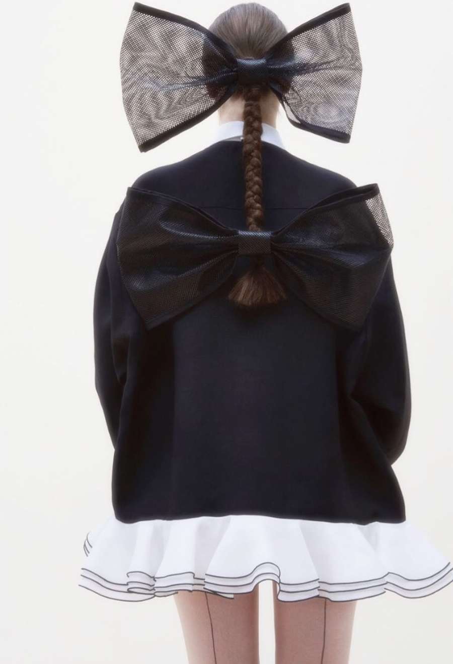 Back view of a woman with a long braided hairstyle, wearing a black top with white ruffled trim and large bow details.