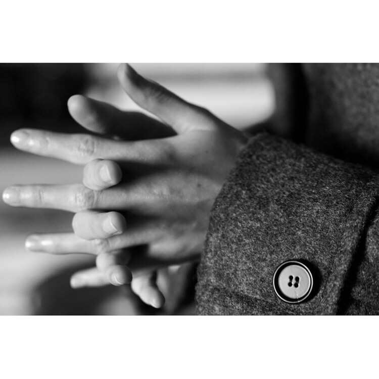Close-up black and white photo of two hands intertwined, wearing a wool coat with a button in a softly lit indoor setting.