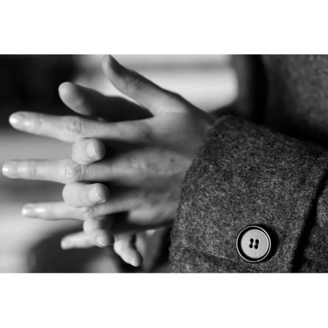 Close-up black and white photo of two hands intertwined, wearing a wool coat with a button in a softly lit indoor setting.