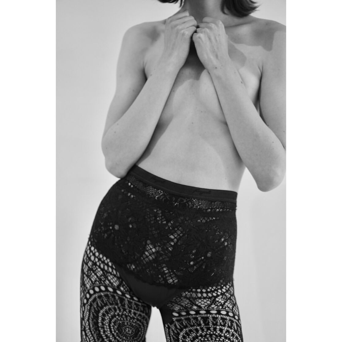 A nude adult female poses in a photo studio wearing lace patterned tights against a white background. Monochrome fashion body portrait.