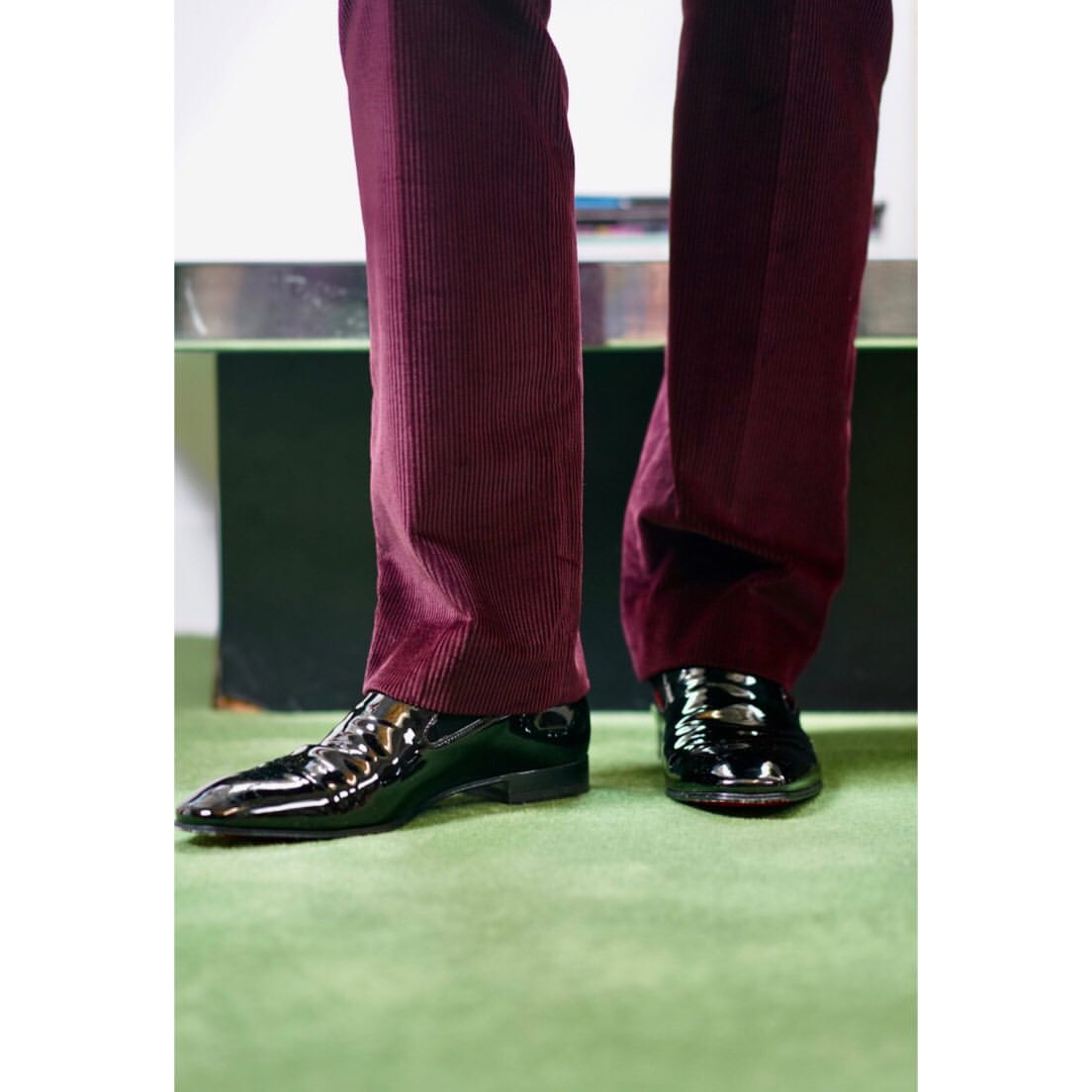 Close-up of glossy black leather dress shoes and dark red corduroy pants, paired with a vibrant green carpet, creating a stylish contrast.