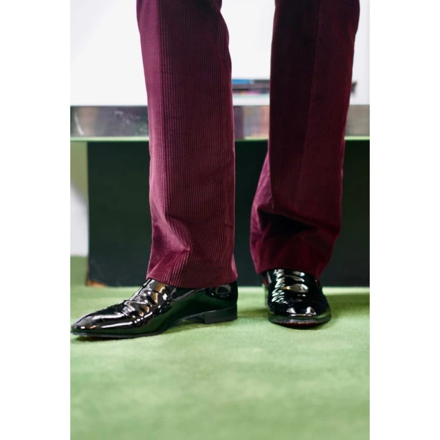 Close-up of glossy black leather dress shoes and dark red corduroy pants, paired with a vibrant green carpet, creating a stylish contrast.