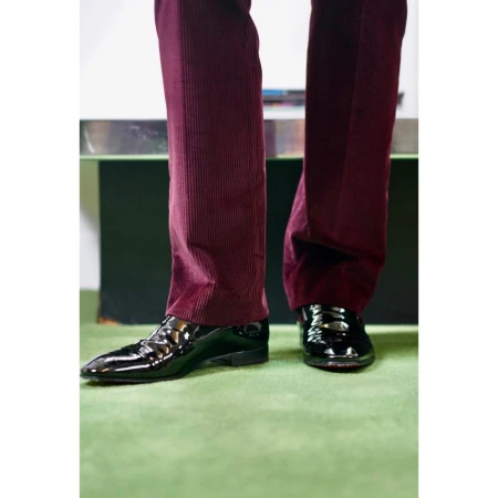 Close-up of glossy black leather dress shoes and dark red corduroy pants, paired with a vibrant green carpet, creating a stylish contrast.