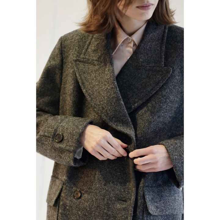 A woman fastens a dark grey, wool double-breasted coat. Her hands are visible at the waist with a soft pink shirt collar showing.