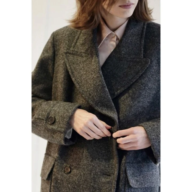 A woman fastens a dark grey, wool double-breasted coat. Her hands are visible at the waist with a soft pink shirt collar showing.