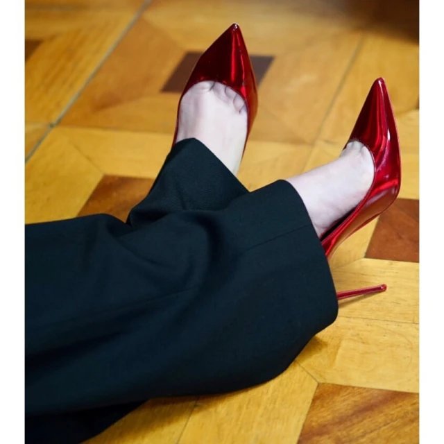 Close-up of pointed-toe heeled shoes in metallic red, paired with dark trousers, on a parquet floor. Fashion footwear.