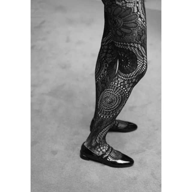 Close-up of a woman's legs and black patent leather shoes wearing patterned black lace tights against a neutral gray background.