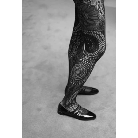 Close-up of a woman's legs and black patent leather shoes wearing patterned black lace tights against a neutral gray background.