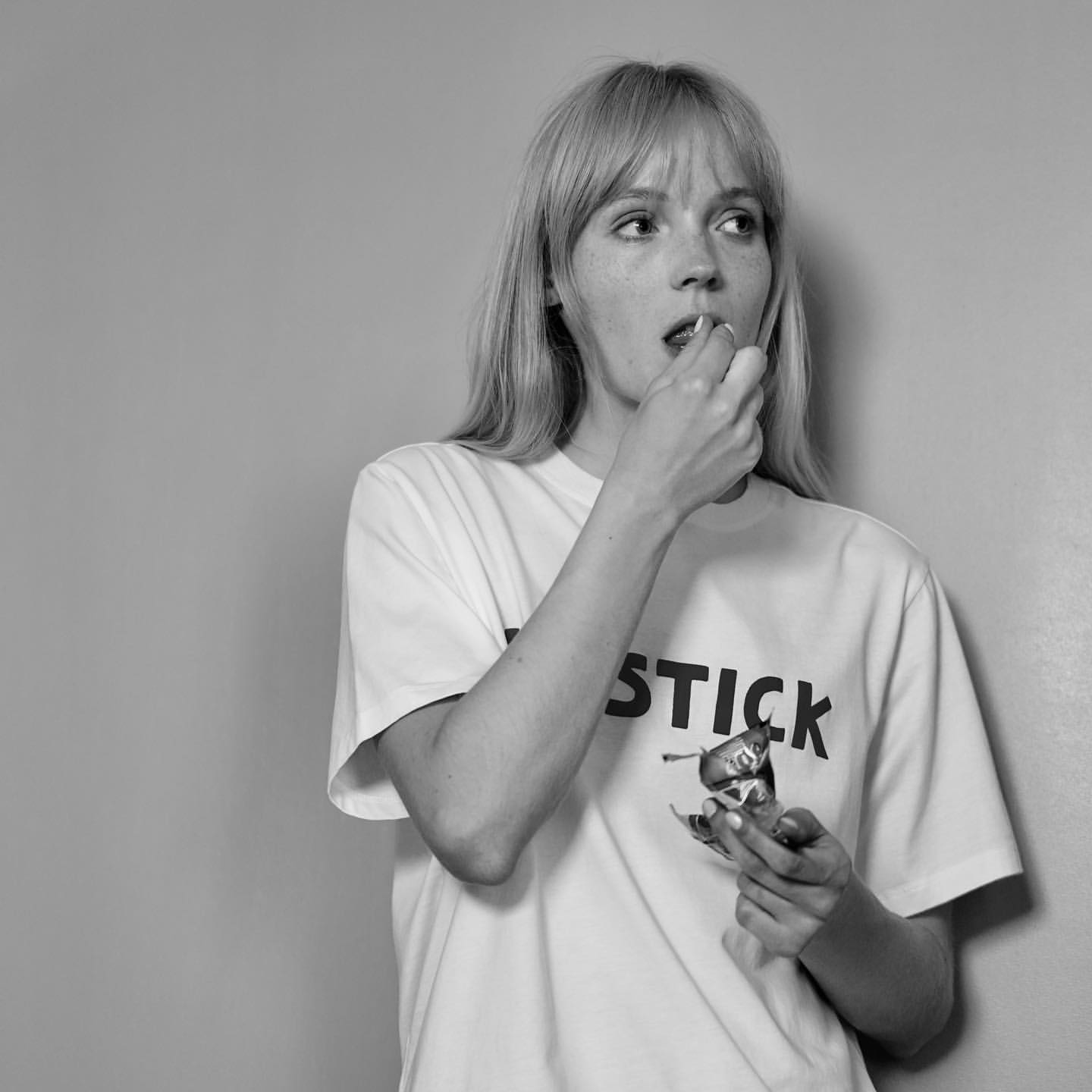 Blonde woman in white tee with black lipstick graphic eats candy. Indoor shot in black and white with a grey backdrop.