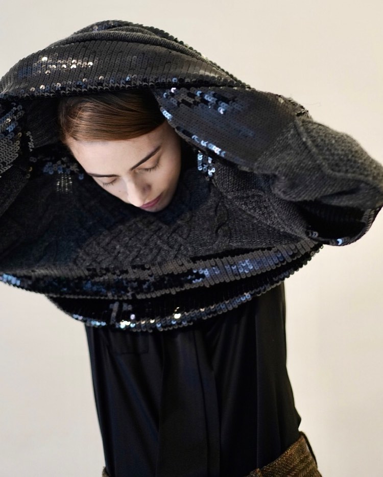 Woman in a black, sequined jacket poses in a studio with her head slightly tilted down. The scene is minimalist and fashion-focused.