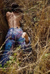 Person lying face down in tall dry golden grass wearing colourful embroidered blue jeans and bare feet