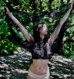 Woman in dark sheer mesh top holding up black fabric standing outdoors in forest by Lynette Garland