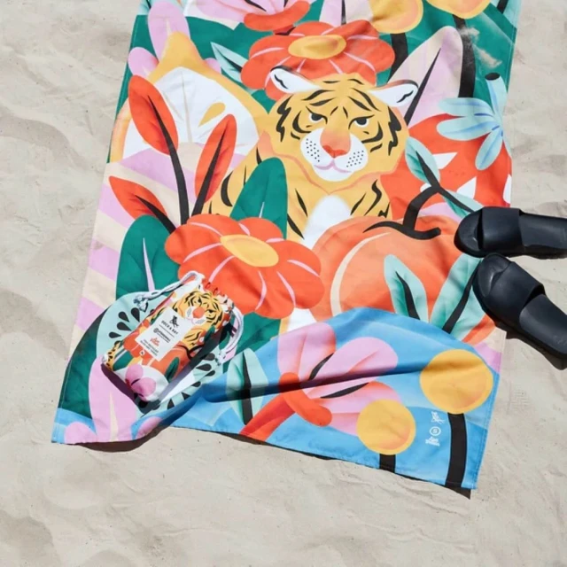 Colorful beach towel with a tiger and floral design laying on the sand next to a matching drawstring bag and a pair of sandals.