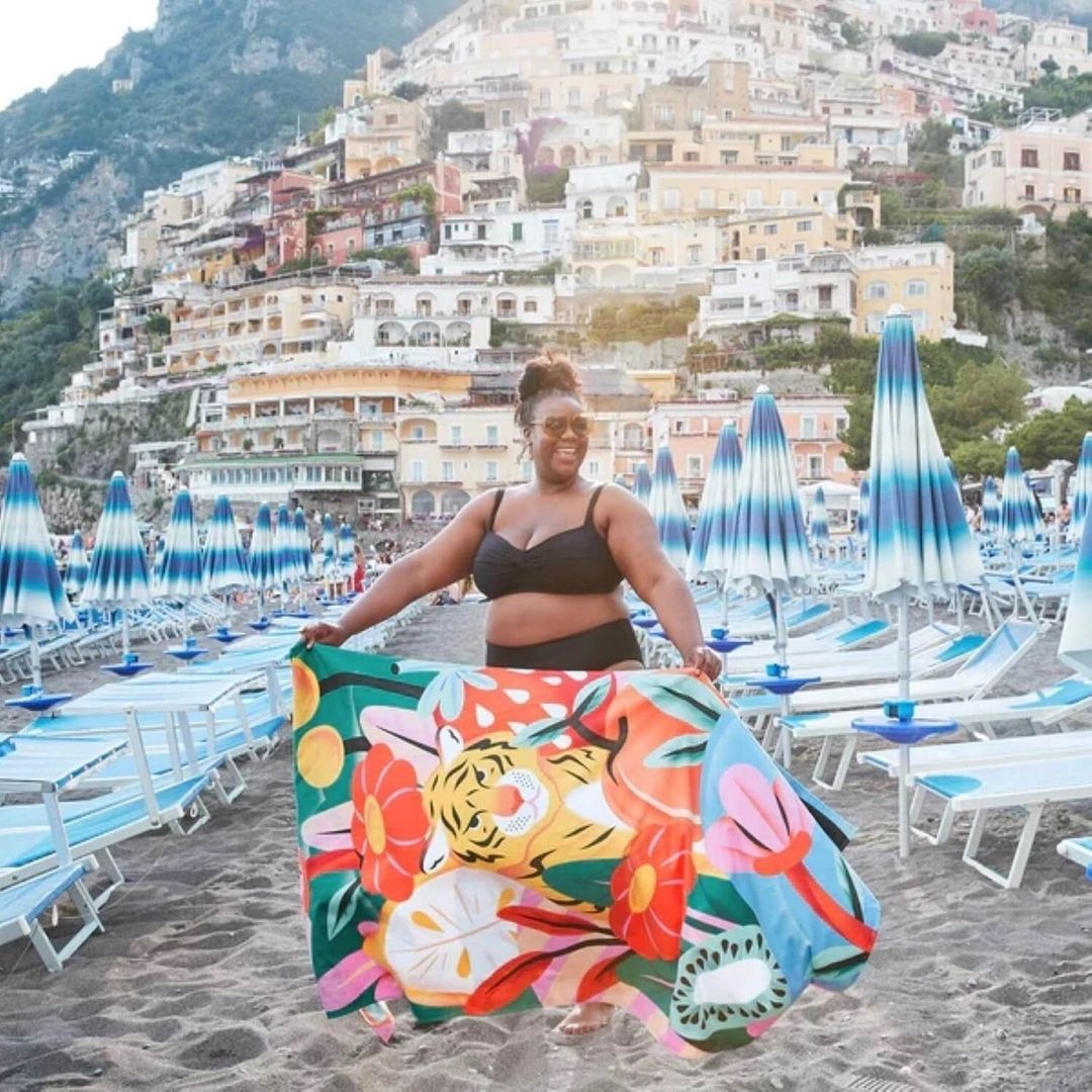 Woman on a beach in a black bikini, holding a colourful graphic beach towel, with blue-and-white striped umbrellas and buildings in the background.