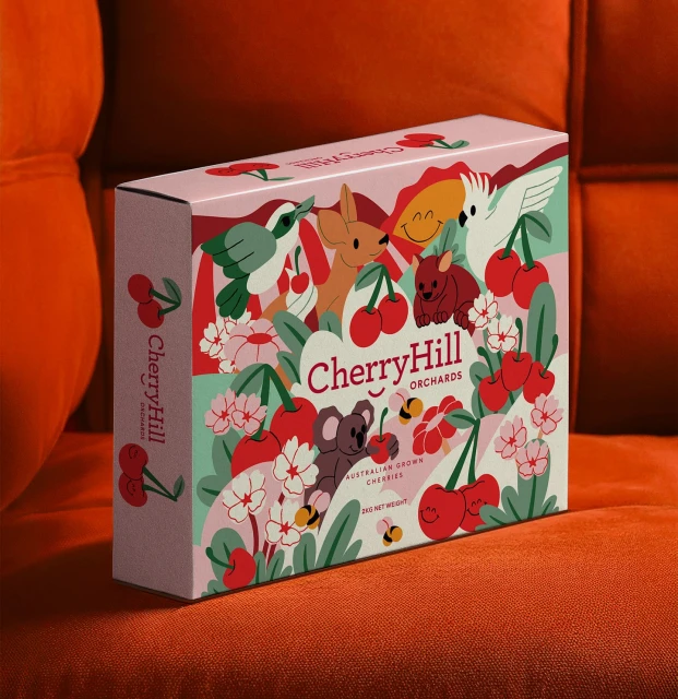 Close-up of a CherryHill Orchards gift box, featuring cartoon animal illustrations and cherries, against an orange couch backdrop.