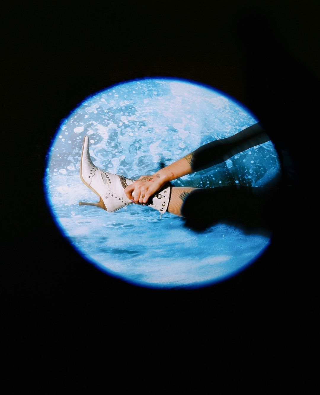An arm and leg are visible in a circle of blue light against a dark background, focusing on a studded high-heeled boot.