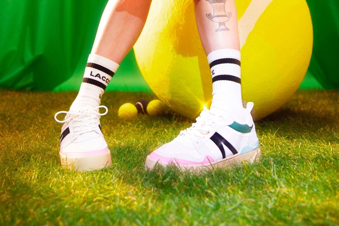 Close-up of Lacoste sneakers on grass with a yellow ball, black and white socks on a woman's legs, and a tattoo of a trophy.