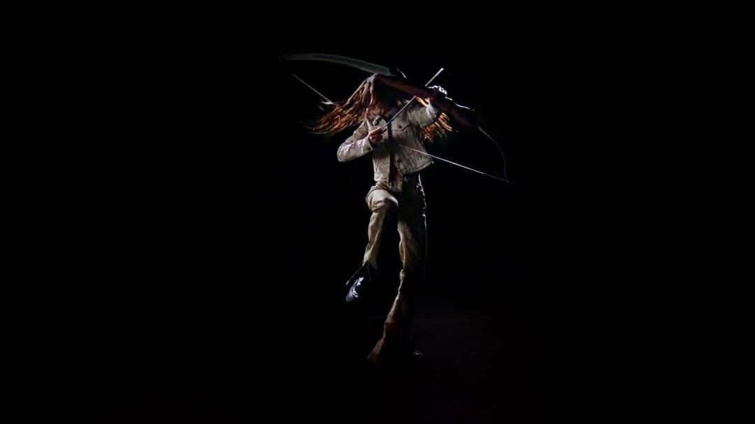 A woman with long brown hair, wearing a white jacket and pants, holds a bow and arrow in a dramatic pose against a black backdrop.