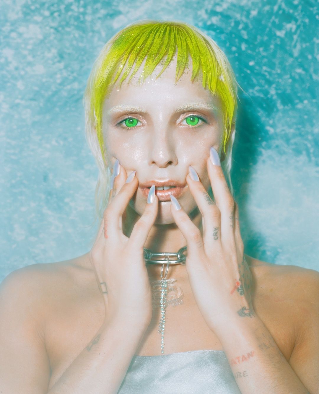 Close-up portrait of a woman with fair skin, neon green hair, and green eyes, posing in front of a soft blue and white textured background.