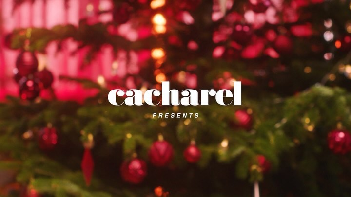 Christmas tree decorated with red ornaments and lights, set against a blurred background, featuring the 'Cacharel' logo and text.