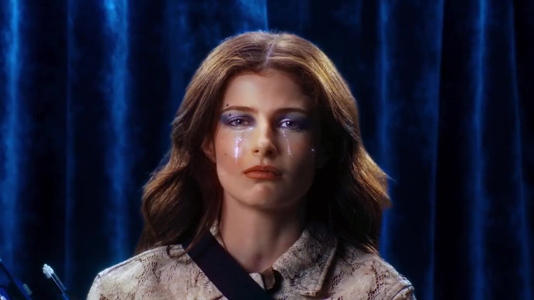 Close-up of a female with brown hair and dramatic makeup with tears. She wears a patterned jacket and stands against a blue backdrop.