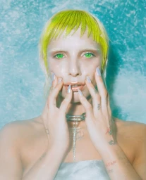 Portrait of a model with neon green hair and bright green eyes holding hands up to face against textured blue background