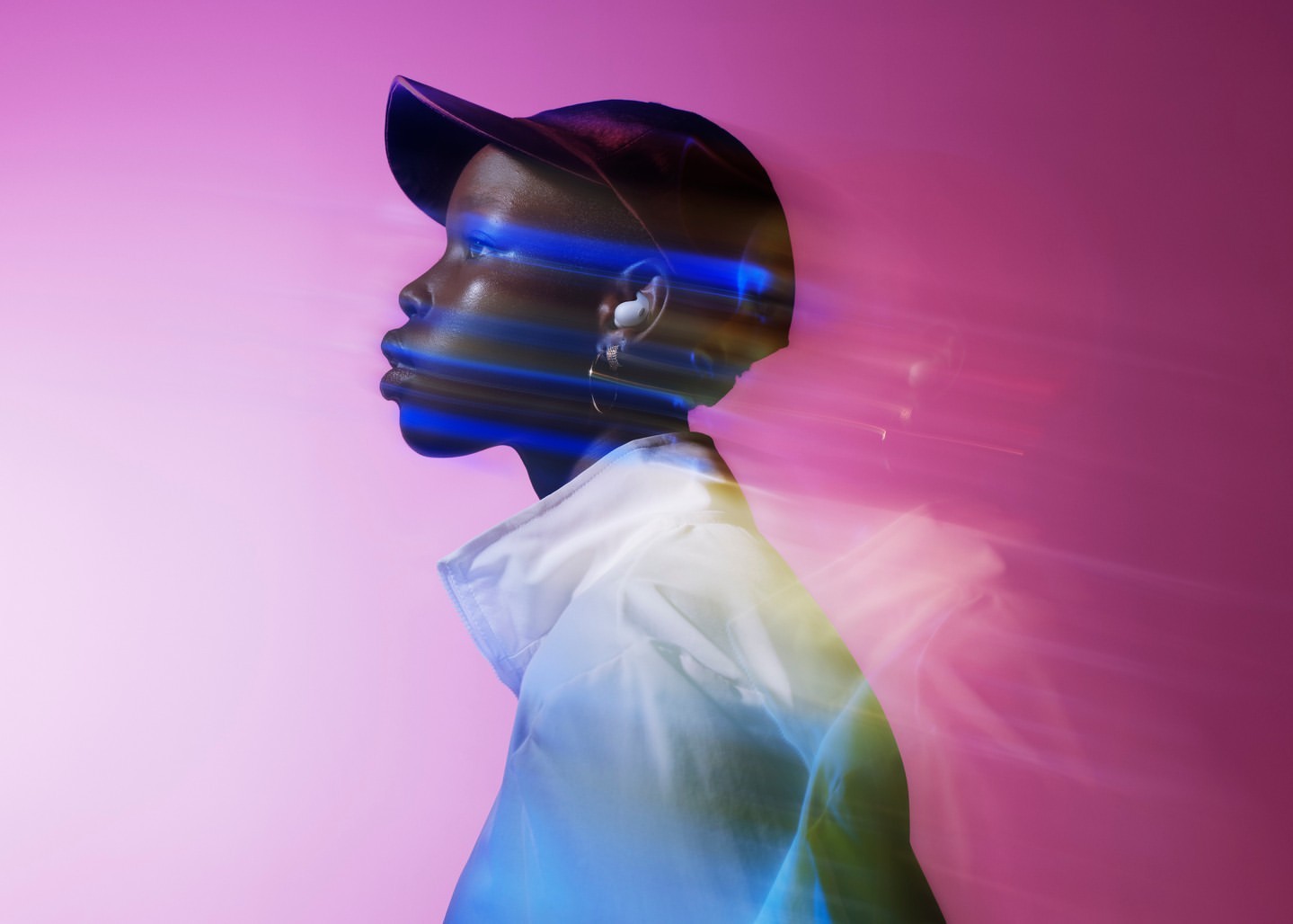 Profile of a Black adult in a white jacket and cap against a pink background with blue and yellow light streaks and earrings.
