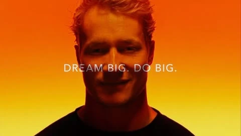 Portrait of fair-skinned adult male in black top with short blonde hair against a vivid gradient orange and yellow background with text overlay.
