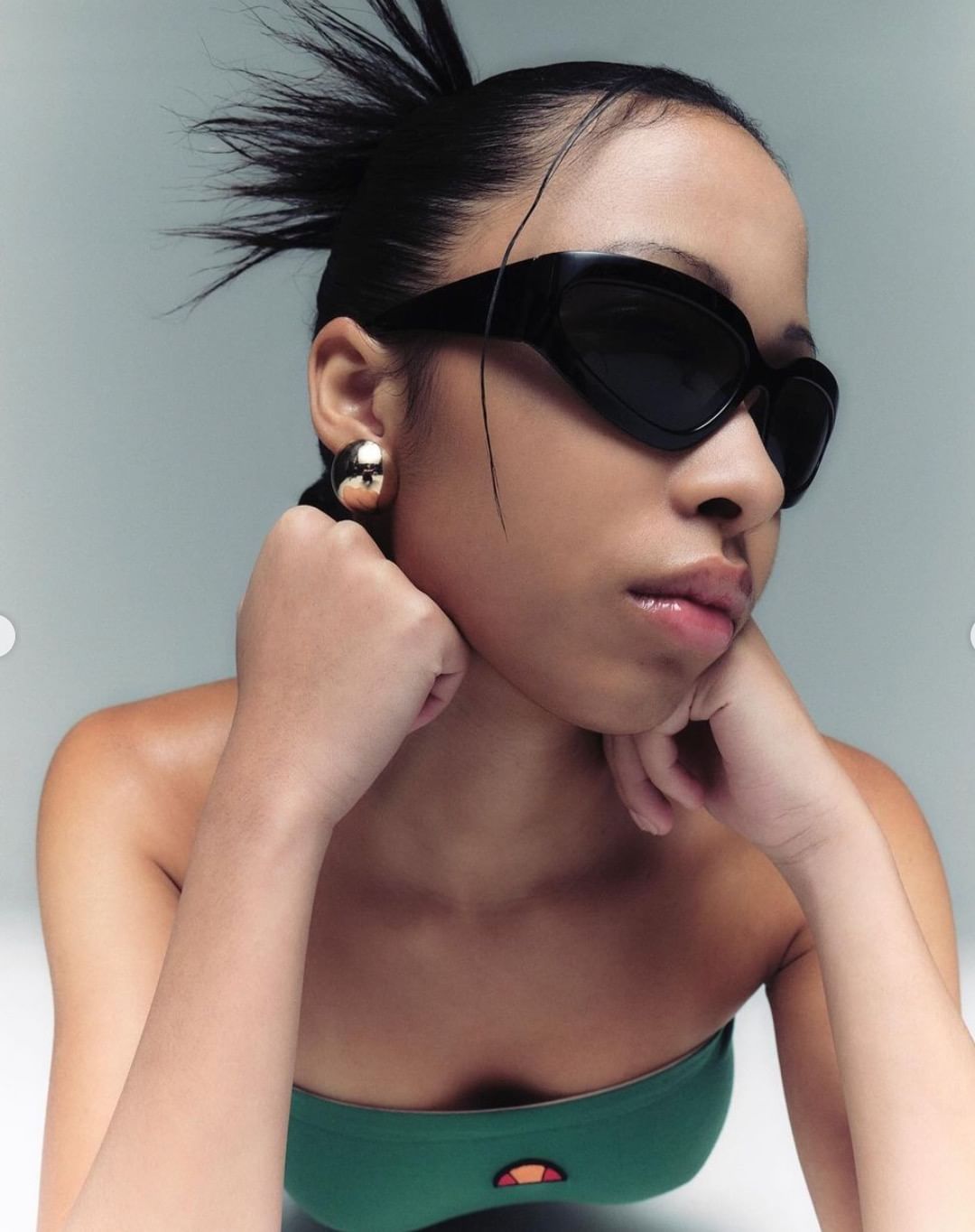 Close-up of a young Asian woman wearing black sunglasses and gold earrings, gazing directly at the viewer in a fashion editorial.