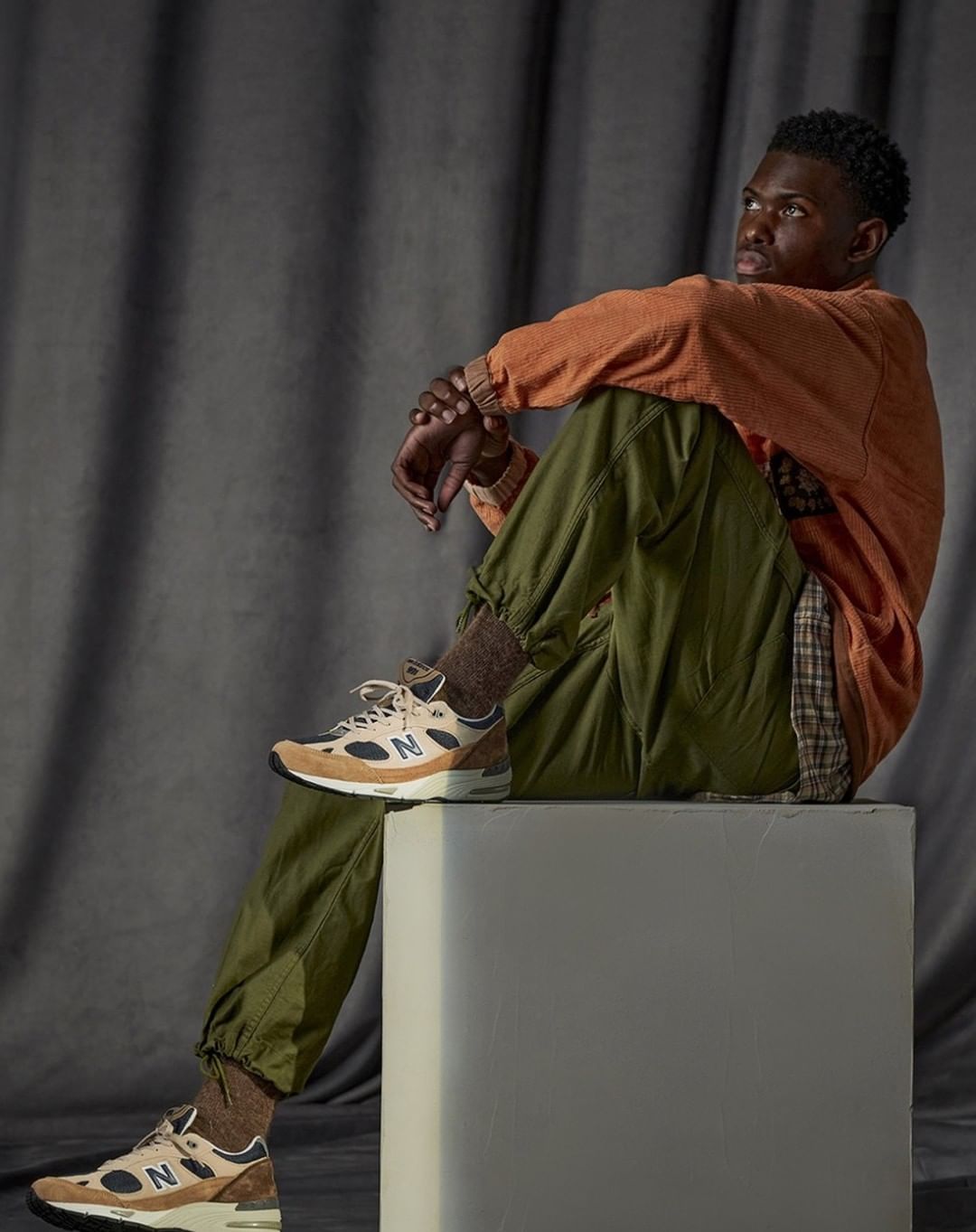 Young Black man seated on a block wearing an orange pullover, green trousers, and sneakers, set against draped fabric.