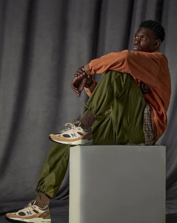 Young Black man seated on a block wearing an orange pullover, green trousers, and sneakers, set against draped fabric.