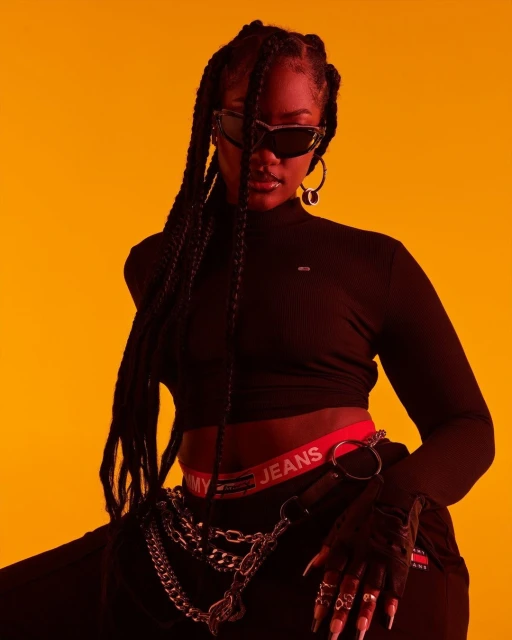 Stylish young woman in a black top, sunglasses, and braids, poses against a bright orange background in a fashion portrait.