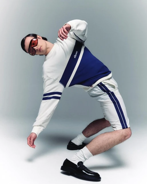 Athletic man in a white-and-navy Ellesse tracksuit and black loafers strikes a dynamic pose in a fashion studio setting.