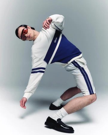 Athletic man in a white-and-navy Ellesse tracksuit and black loafers strikes a dynamic pose in a fashion studio setting.