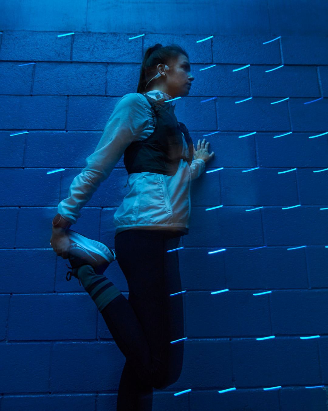 Athletic woman in activewear stretches quadriceps against blue textured wall with neon lights, preparing for a workout, fitness scene.