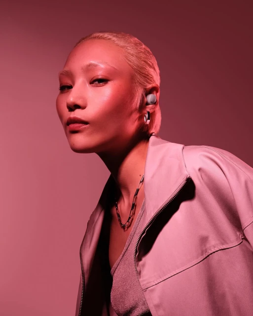 Close-up studio shot of a stylish adult woman with short blonde hair wearing wireless earbuds, a light jacket, and a necklace, posing.