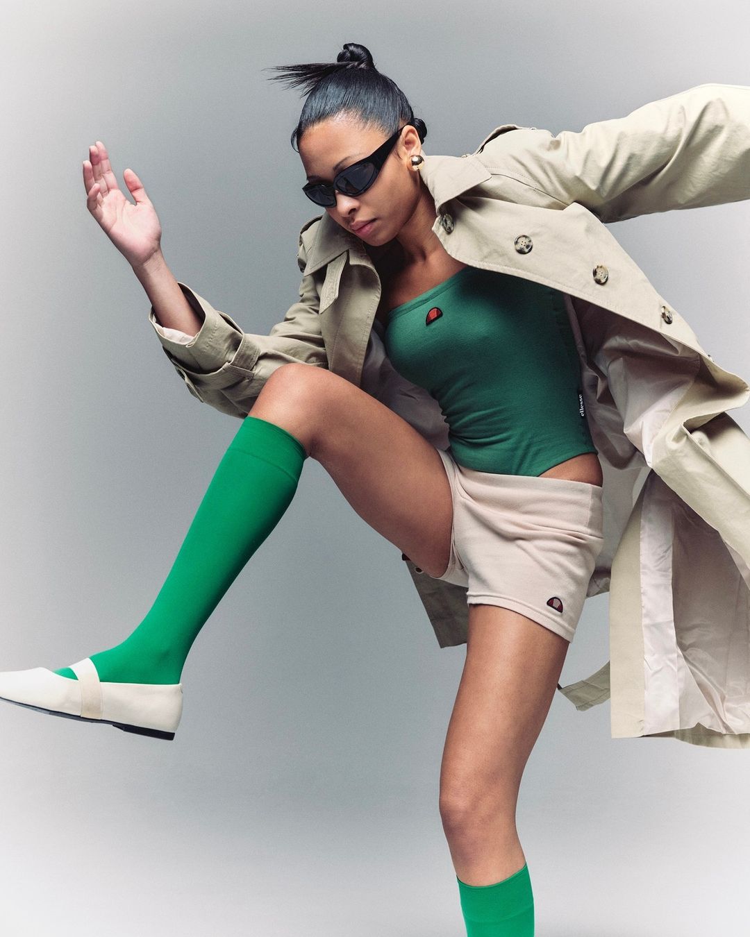 A woman strikes a pose in a green bodysuit, beige shorts, a trench coat, and green knee socks against a light background.