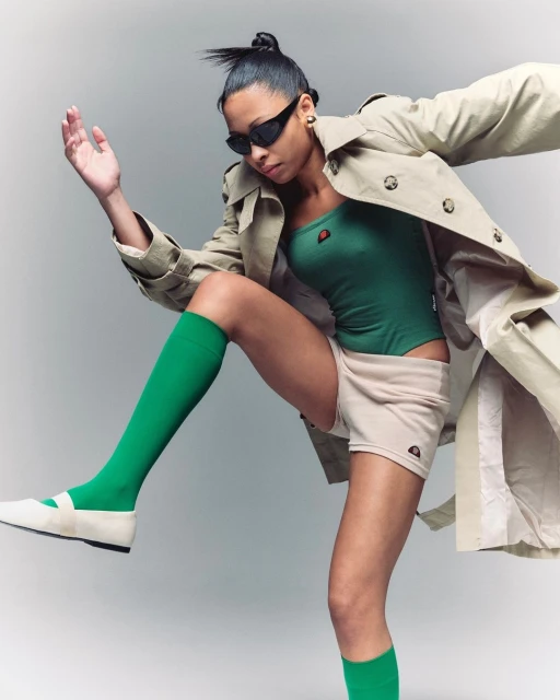 A woman strikes a pose in a green bodysuit, beige shorts, a trench coat, and green knee socks against a light background.