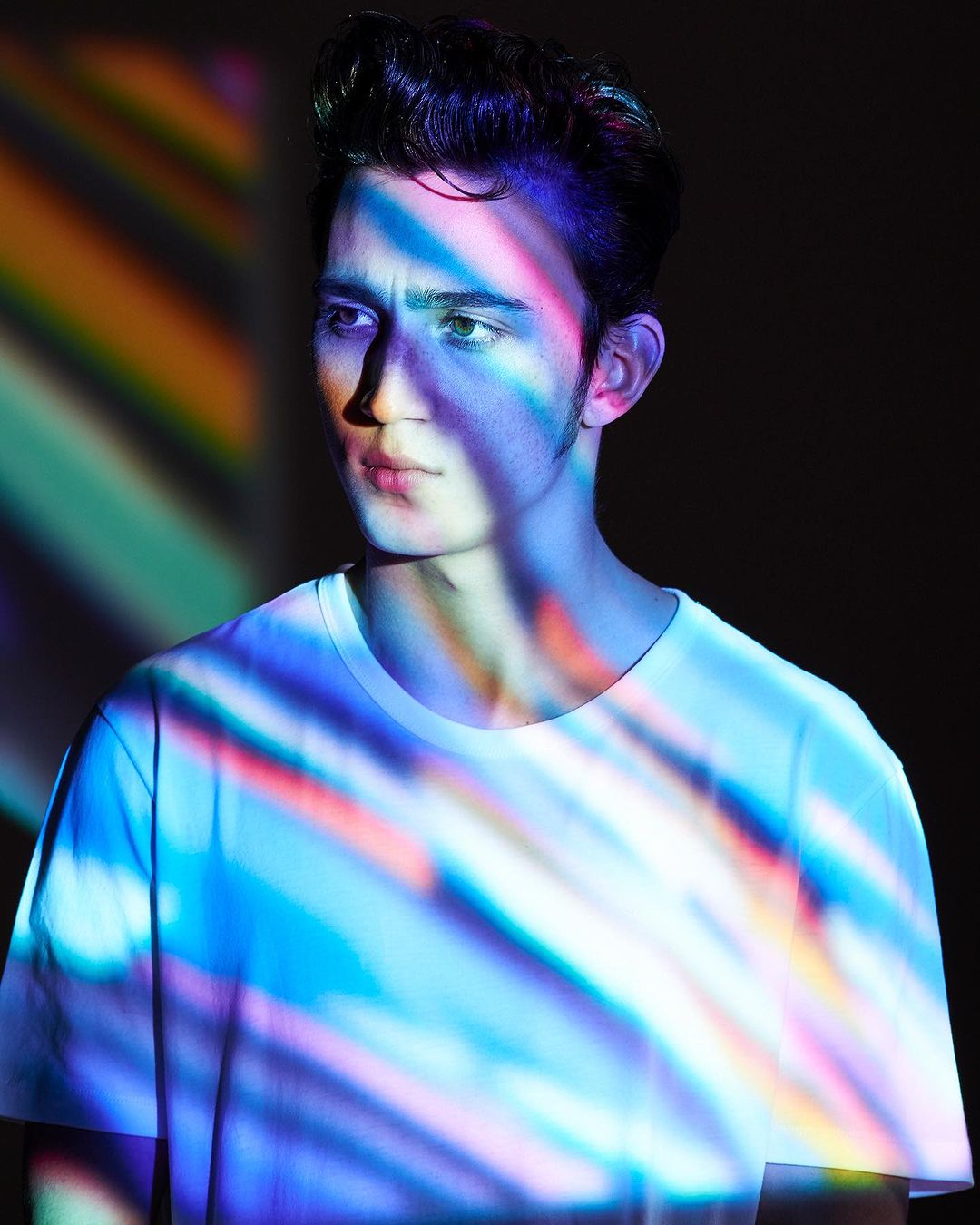 Portrait of a young man with dark hair and a white t-shirt, illuminated by dynamic multicolored lighting in a dark indoor setting.