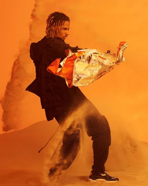 Adult male model poses in a sand-filled studio with orange lighting, holding reflective material while wearing a black jacket and pants.